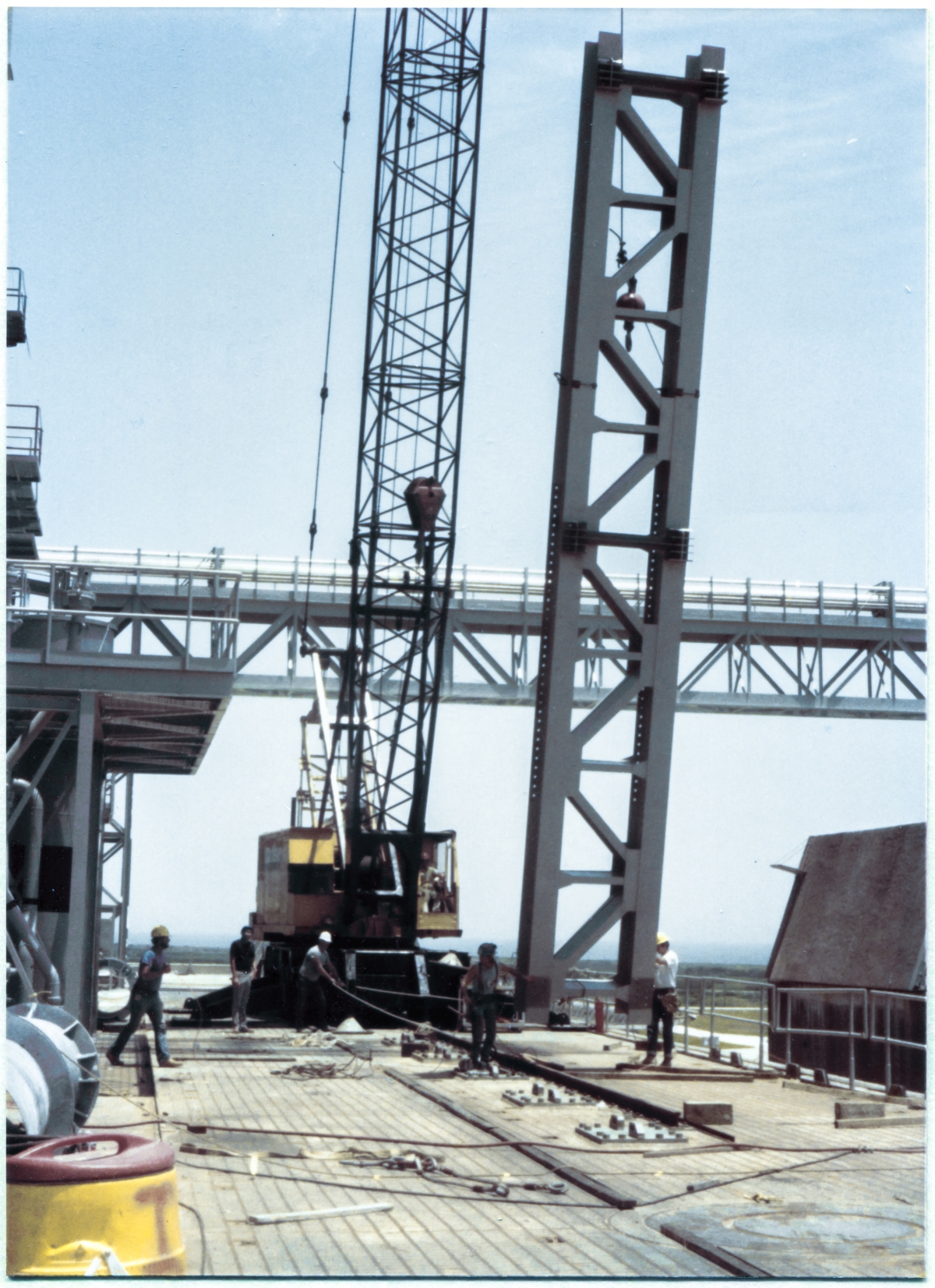 Image 072. The GOX Arm Strongback lift is now fully under way, with the strongback clear of the ground which was supporting it, now hanging in suspension, supported solely by its outré lifting sling which is attached via a set of shackles up above the headache ball and the crane's lifting hook beneath that. In this image, the headache ball can clearly be seen hanging off-center, away from the crane's jib line, its own attach hardware pulled to the side at a significant angle, bearing no load above and beyond its own weight. From this angle, the choker slings wrapped around the twin W24x104's which constitute the main body of the Strongback can be seen resting on pads at each corner of the wide-flanges to keep from chafing, possibly damaging not only the Strongback, but also the wire ropes of the slings. Photo by James MacLaren.